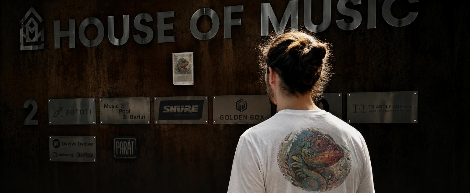 The musician behind Slumbering Shaman, seen from behind, wearing a shirt printed with the Dakota’s Journey chameleon