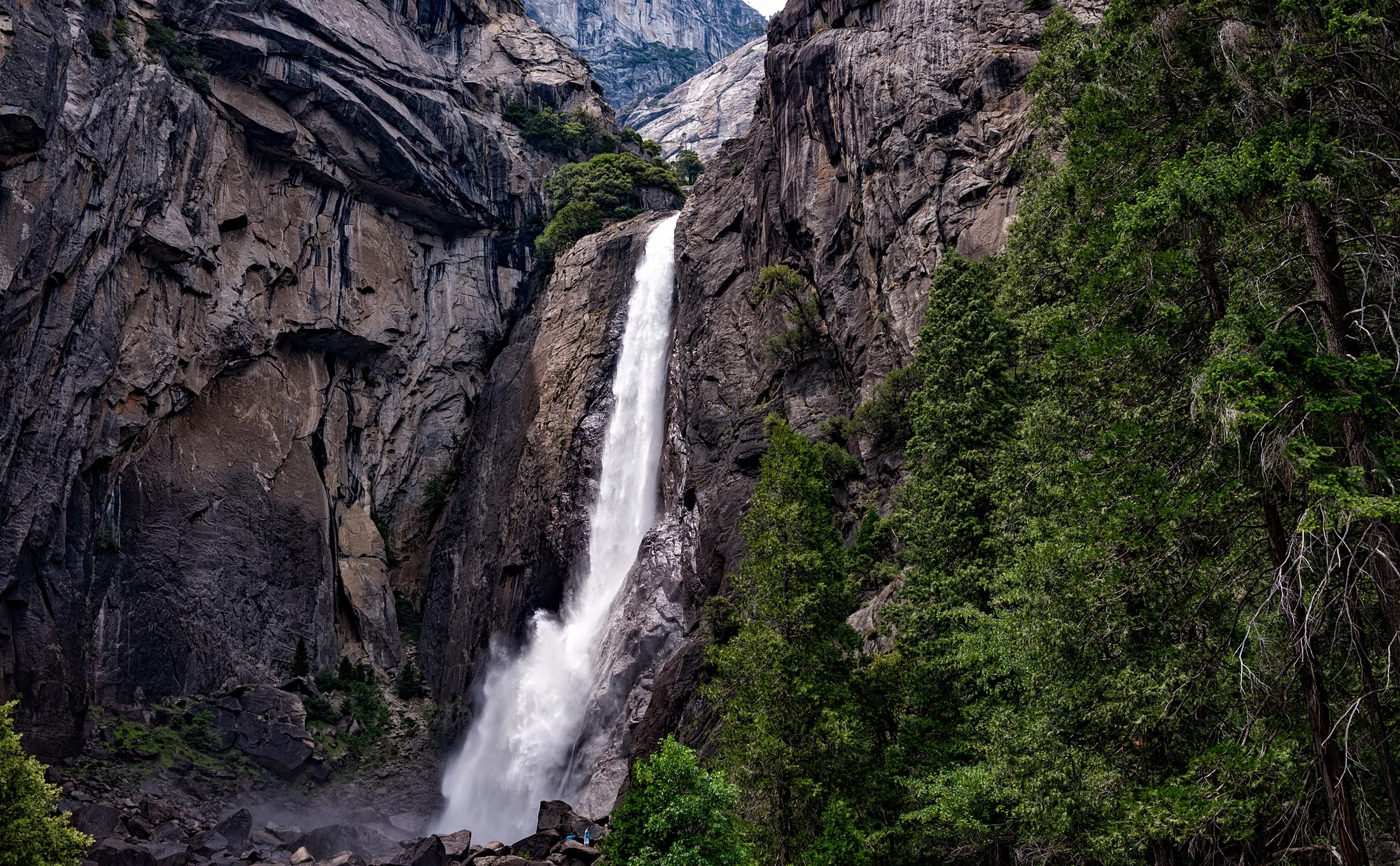 A tall waterfall dropping through a granite gorge, framed by rock walls and dense evergreen trees